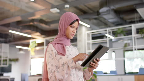 Happy-biracial-casual-businesswoman-in-hijab-using-tablet-in-office,-slow-motion