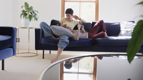 Happy-caucasian-lesbian-couple-lying-on-sofa,-using-tablet-and-smartphone-and-smiling-in-sunny-house