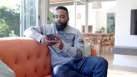 Afroamerikanischer-Mann-Benutzt-Smartphone-Im-Sonnigen-Wohnzimmer,-Zeitlupe