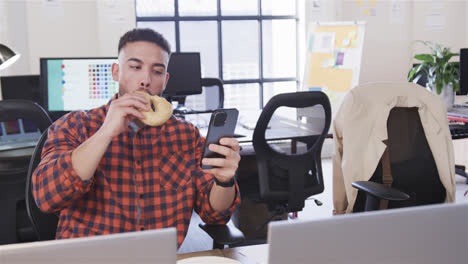 Biracial-male-sitting-at-desk,-eating,-looking-at-smartphone-and-smiling,-slow-motion