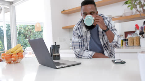 Hombre-Afroamericano-Haciendo-Café-Y-Usando-Una-Computadora-Portátil-En-Una-Cocina-Soleada,-Cámara-Lenta