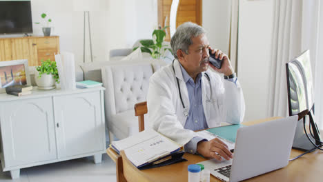 Senior-biracial-male-doctor-sitting-at-desk-talking-on-smartphone-and-using-laptop