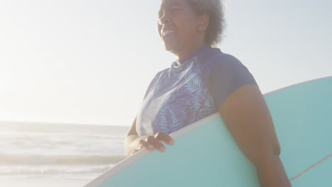 Glückliche-ältere-Afroamerikanische-Frau,-Die-In-Zeitlupe-Am-Strand-Spazieren-Geht-Und-Ein-Surfbrett-Hält