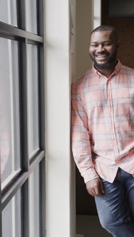 Vertical-video-portrait-of-happy-african-american-casual-businessman-standing-by-window-in-office