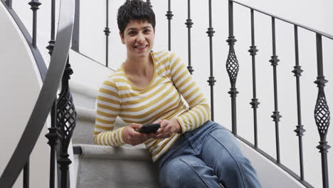 Happy-caucasian-woman-sitting-on-stairs,-holding-smartphone-and-smiling-in-sunny-house
