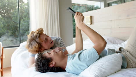 Happy-diverse-gay-male-couple-lying-on-bed-and-using-smartphone-at-home,-slow-motion