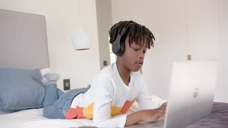 Glücklicher-Afroamerikanischer-Junge-Mit-Kopfhörern,-Der-Zu-Hause-Mit-Einem-Laptop-Auf-Dem-Bett-Liegt,-Zeitlupe