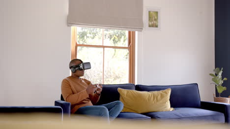 African-american-man-sitting-on-sofa-using-vr-headset-and-playing-video-games-at-home,-slow-motion