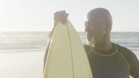 Glücklicher-älterer-Afroamerikanischer-Mann-Mit-Surfbrett-Am-Strand,-In-Zeitlupe