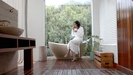 Biracial-woman-wearing-bathrobe-sitting-on-bathtub-drinking-tea-in-bathroom-at-home,-slow-motion