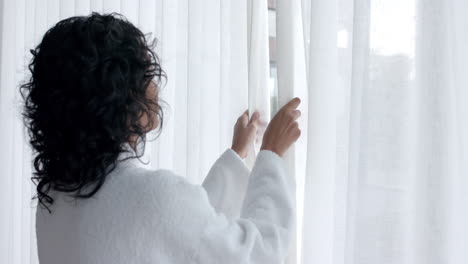 Biracial-woman-wearing-bathrobe-opening-curtains-and-looking-through-window-at-home,-slow-motion