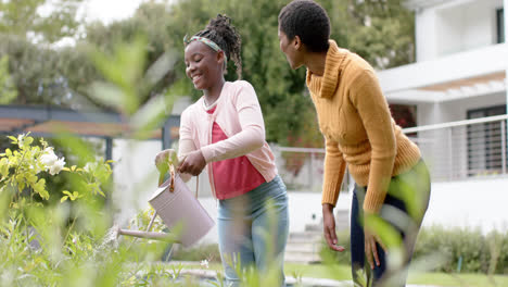 Glückliche-Afroamerikanische-Tochter-Und-Mutter-Gießen-Pflanzen-Im-Garten,-Zeitlupe