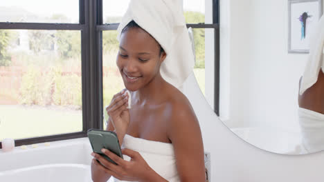 Happy-biracial-woman-with-towel-on-head-washing-her-face-and-using-smartphone,-slow-motion