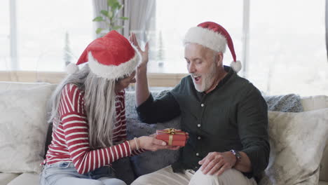 Middle-aged-caucasian-couple-in-santa-hats-gifting-at-christmas-at-home,-slow-motion