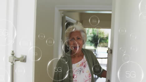 Animation-Eines-5G-Textes-In-Kreisen-über-Einer-Lächelnden-älteren-Frau-Mit-Gemischter-Abstammung,-Die-Die-Tür-Eines-Hauses-öffnet