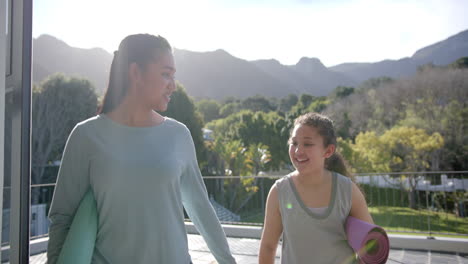 Happy-biracial-mother-and-daughter-walking-with-yoga-mats-on-terrace-in-sunny-day,-slow-motion