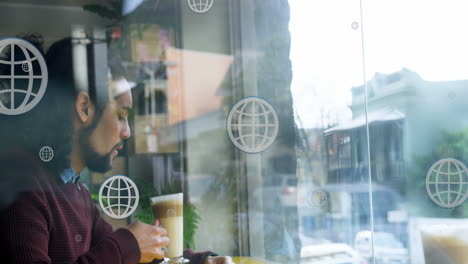 Animation-of-globe-icons-over-biracial-man-drinking-coffee-while-looking-through-glass-window