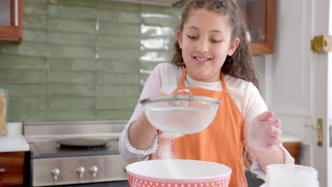 Happy-biracial-girl-with-long,-curly-hair-sifting-flour-and-smiling-in-sunny-kitchen