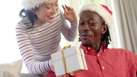 Happy-diverse-couple-in-christmas-hats-exchanging-gift-in-sunny-living-room,-slow-motion