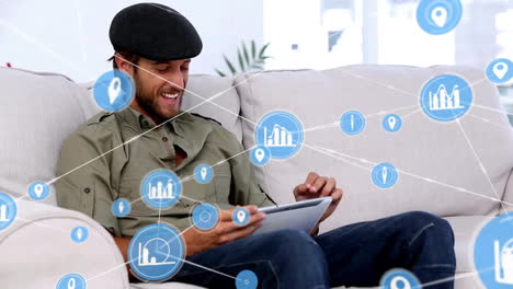 Animation-of-connected-icons-over-caucasian-man-sitting-on-sofa-scrolling-on-digital-tablet-at-home