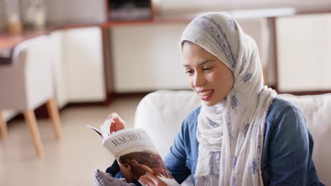 Feliz-Mujer-Birracial-En-Hijab-Leyendo-Un-Libro-En-Un-Sofá-En-Casa-Con-Espacio-Para-Copiar,-Cámara-Lenta