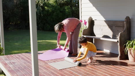 Senior-biracial-grandmother-and-grandson-rolling-yoga-mats-on-terrace,-slow-motion