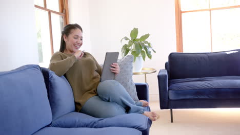Happy-biracial-woman-sitting-on-sofa,-smiling-and-using-tablet-in-sunny-living-room