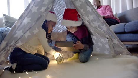 Happy-african-american-brother-and-sister-in-christmas-hats-with-tablet-in-blanket-tent,-slow-motion
