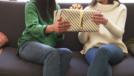 Midsection-of-biracial-mother-and-adult-daughter-exchanging-christmas-gift-on-couch,-slow-motion
