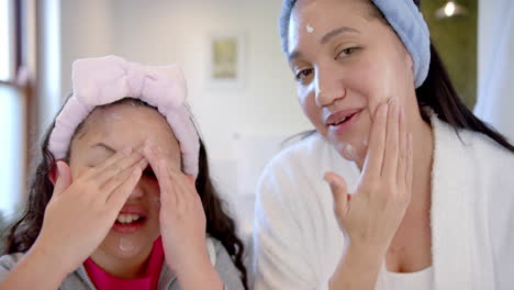 Glückliche-Biracial-Mutter-Und-Tochter-Mit-Stirnbändern-Creme-Auf-Gesichter-In-Sonnigen-Badezimmer