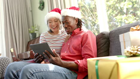 Happy-diverse-couple-in-christmas-hats-using-tablet-in-sunny-living-room,-slow-motion