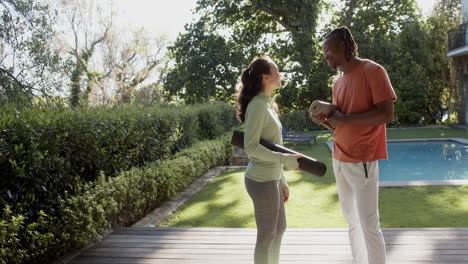 Happy-diverse-couple-holding-yoga-mats-talking-in-sunny-garden,-copy-space,-slow-motion