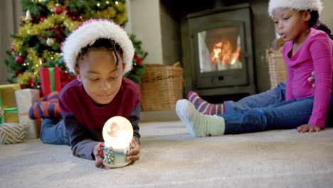 Afroamerikanischer-Sohn-In-Weihnachtsmütze-Hält-Leuchtende-Schneekugel,-Familie-Im-Hintergrund,-Zeitlupe