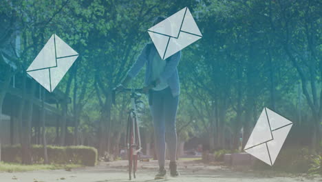 Animation-of-falling-envelope-icons-over-biracial-woman-wearing-mask-walking-with-bicycle-in-park