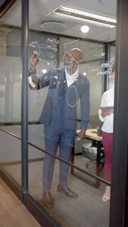 Vertical-video-of-african-american-businessman-writing-on-glass-wall-in-office-with-copy-space