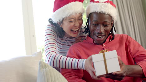 Glückliches,-Vielfältiges-Paar-In-Weihnachtsmützen-Tauscht-Geschenke-Im-Sonnigen-Wohnzimmer-Aus,-Zeitlupe