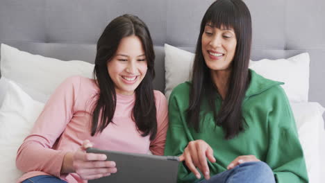 Happy-biracial-mother-and-adult-daughter-using-tablet-sitting-on-bed,-slow-motion