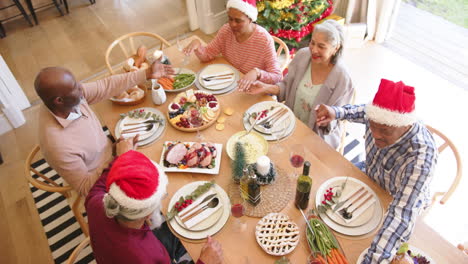 Happy-diverse-senior-friends-holding-hands-to-say-grace-at-christmas-dining-table,-slow-motion