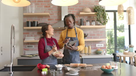 Happy-diverse-couple-in-aprons-using-tablet-and-baking-in-kitchen,-copy-space,-slow-motion