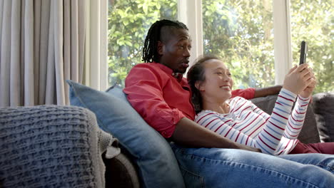 Happy-diverse-couple-relaxing-on-couch-using-tablet-in-living-room,-copy-space,-slow-motion