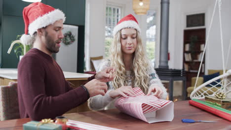 Happy-diverse-couple-sitting-at-table-packing-christmas-presents-at-home,-in-slow-motion