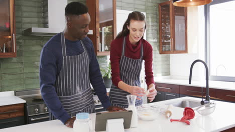 Happy-diverse-couple-standing-in-kitchen,-using-tablet-and-preparing-dough,slow-motion