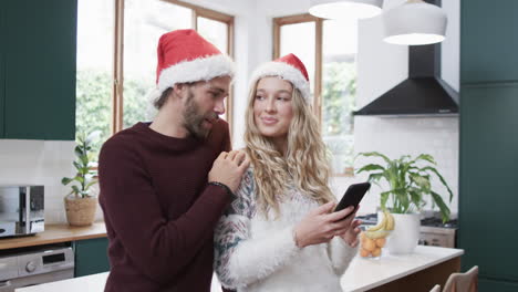 Diverse-couple-wearing-santa-hats-using-smartphone-in-kitchen,-in-slow-motion