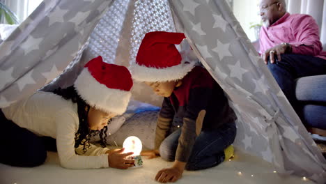 African-american-brother-and-sister-in-christmas-hats-using-snow-globe-in-blanket-tent,-slow-motion
