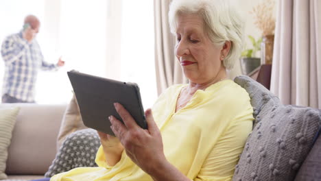 Glückliches,-Vielfältiges-Seniorenpaar-Mit-Smartphone-Und-Tablet-Im-Sonnigen-Wohnzimmer,-Zeitlupe