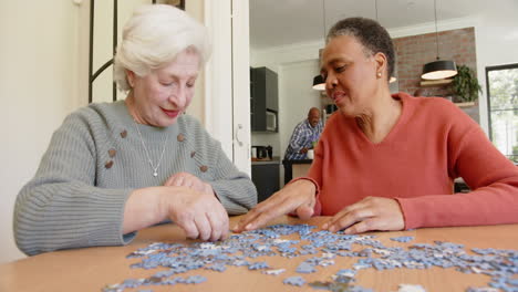 Happy-diverse-senior-female-friends-talking-and-doing-jigsaw-puzzle-at-dining-table,-slow-motion