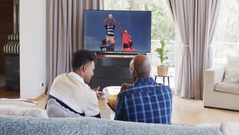 Rear-view-of-african-american-father-and-adult-son-eating-popcorn-watching-rugby-on-tv,-slow-motion