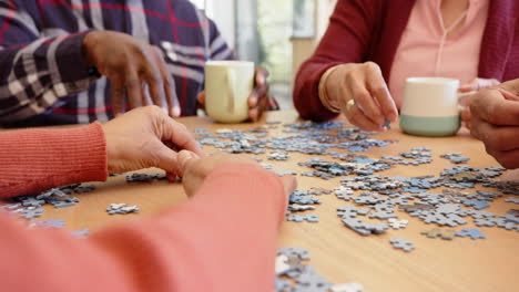 Midsection-of-diverse-senior-friends-having-coffee-doing-jigsaw-puzzle-at-dining-table,-slow-motion