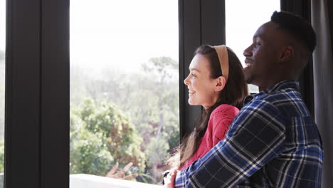 Happy-diverse-couple-looking-out-window,-smiling-and-embracing,-slow-motion,-copy-space