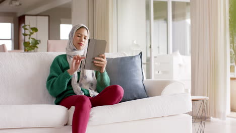 Biracial-woman-in-hijab-using-tablet-on-sofa-at-home-with-copy-space,-slow-motion